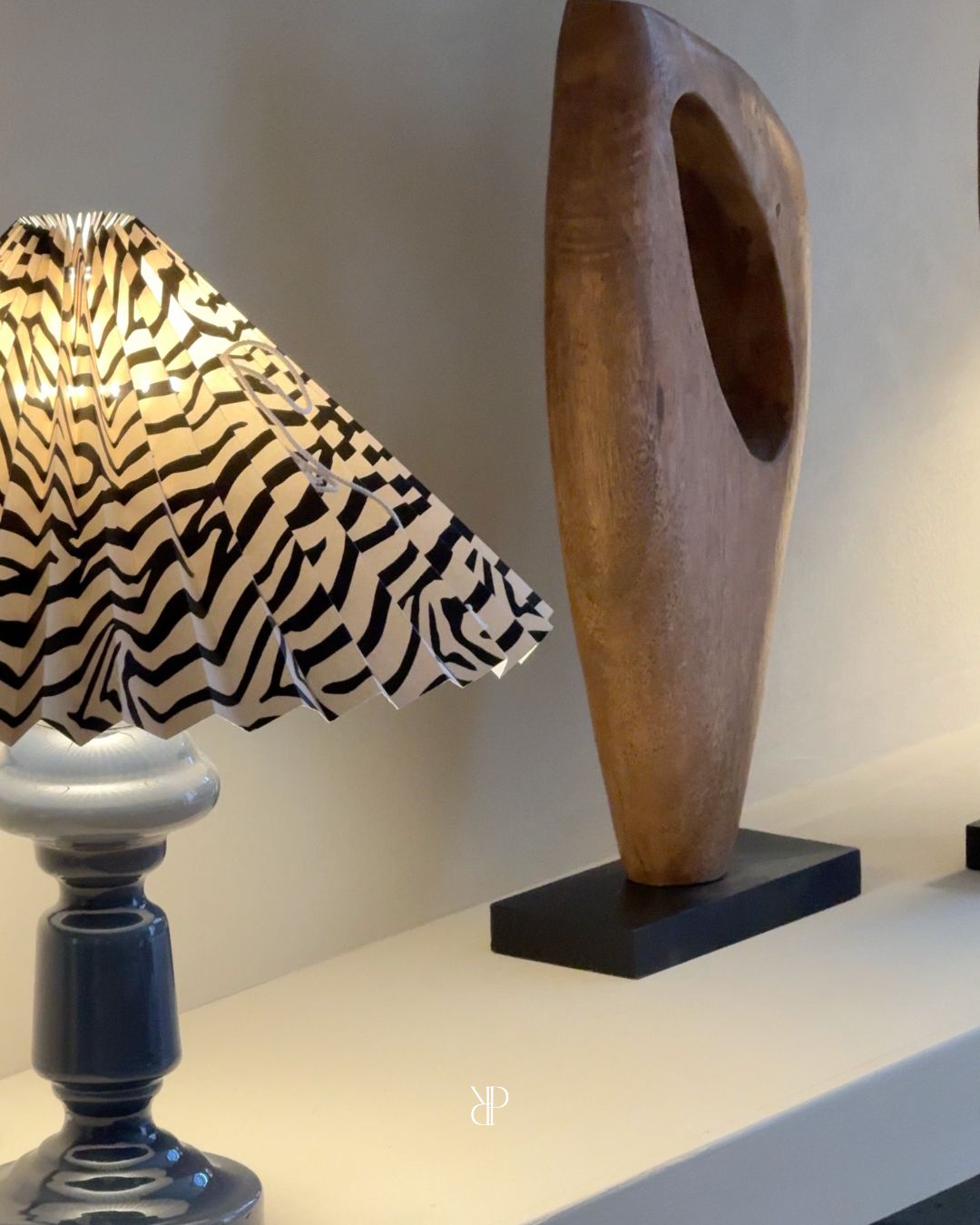 Tændt lampe med zebramønstret plisseret skærm ved siden af en skulptur i træ